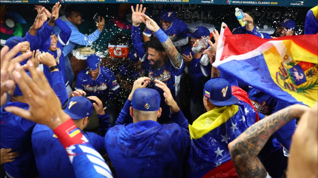 The Venezuelan baseball team at the 2026 World Baseball Classic celebrate their win by joyfully dancing in a circle, waving Venezuelan flags, and throwing sunflower seeds. Everyone looks elated.